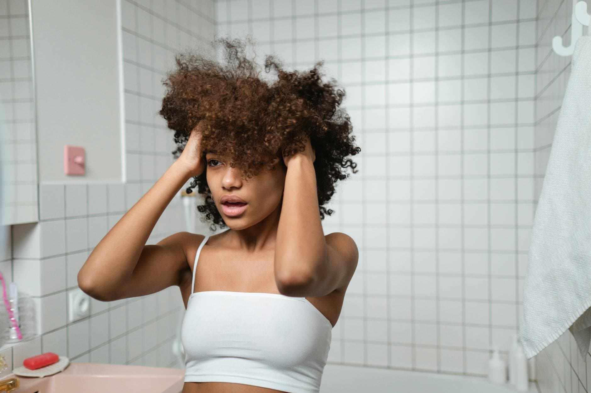 A Black woman with beautiful natural hair applying a hair treatment in a well-lit bathroom, showcasing the pre-pooing process as part of her hair care routine.